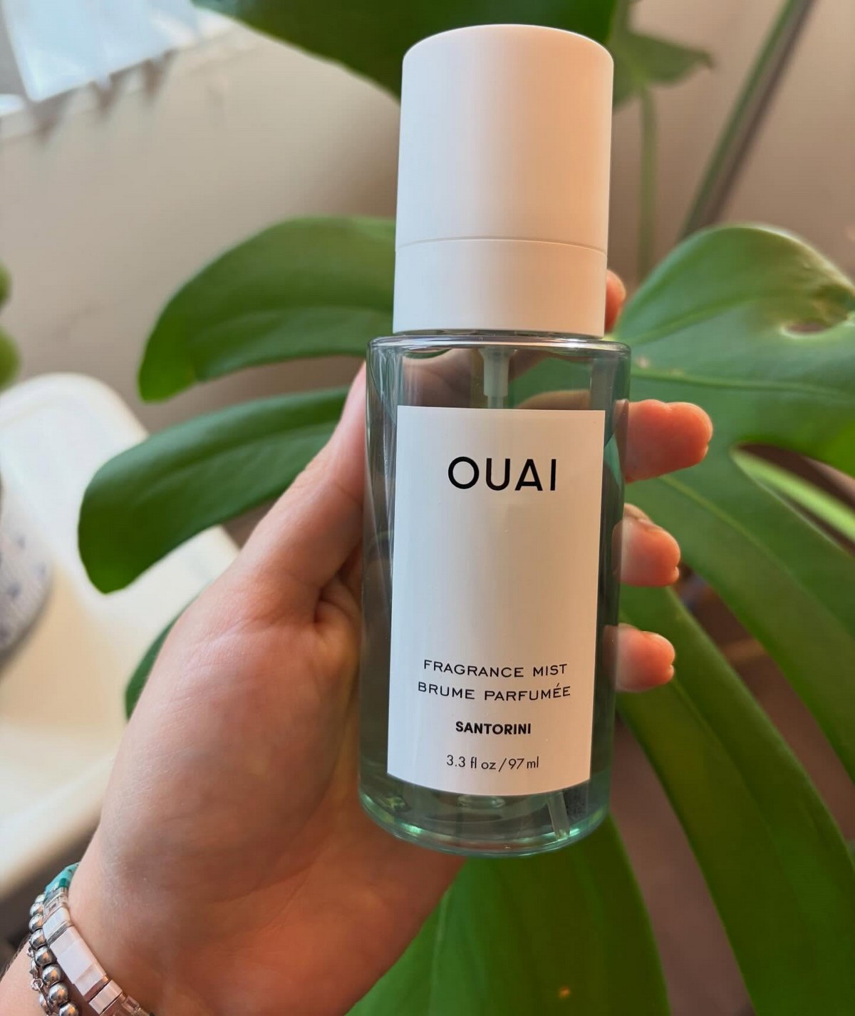 Флакон OUAI Santorini Fragrance Mist у руці на тлі зеленого листя
