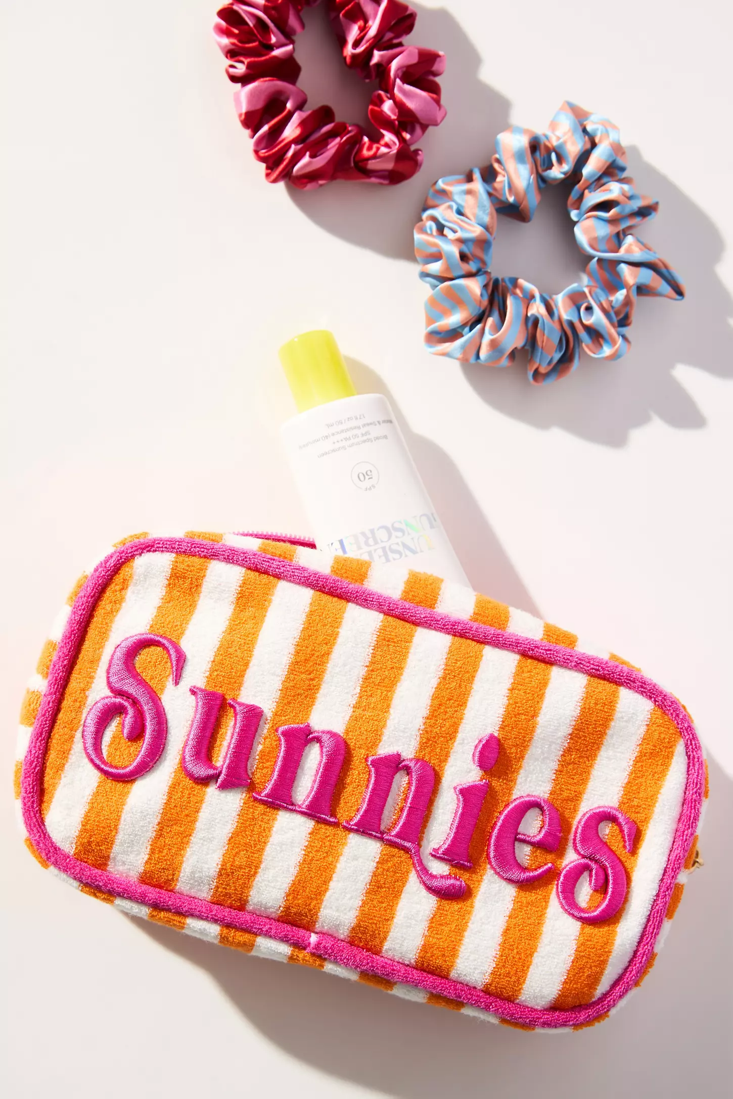 Помаранчево-біла махрова косметичка з написом Sunnies поруч із сонцезахистом і резинками для волосся